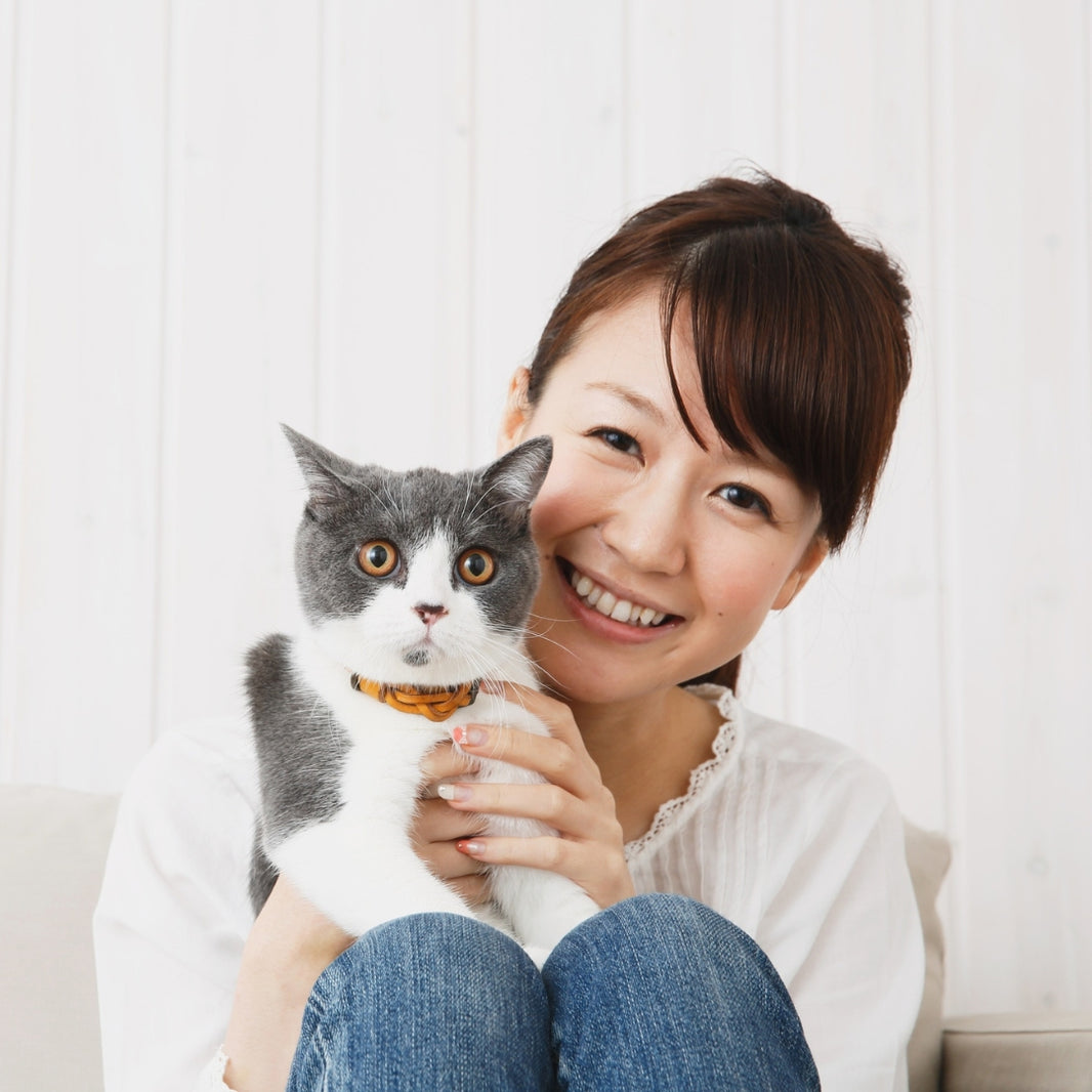 A smiling woman with short brown hair holds a gray and white cat wearing an orange collar, posed against a light white paneled wall.